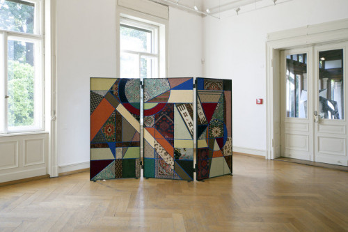 
     <i>Paravent, Social Fabric #3</i>, 
     2013<br />
     Collage with carpets of different origin and production conditions. From knotted kelims, virgin wool and silk carpets to industrially manufactured Tretford, Sisal and wool carpets, 
      180 x 245 x 4 cm<br />
     The subject of borders and transgressing borders is here not just addressed in terms of content, but also formally. The framework of this order are the rational and strict forms of Bauhaus/de Stijl. Nevin Aladağ stages the carpet, one usually passes on foot, vertically, as a pane that constitutes a space – that of the paravent or folding screen. The folding screen serves as protection: originally as wind barrier in China; in western countries it protects the private and intimate from the gaze of strangers. The initially horizontal form of the carpet is brought into a vertical position by the artist, thus blurring the categories of picture and sculpture and changing how they are read. A top view becomes a horizontal view, and we can only read the entire image by walking through the individual motifs. Aladağ here runs through the various connotations of ‘social fabric’: social spaces are constituted by social fabrics, i.e. the demographic, historical, and cultural segments that constitute the structure of a society. This form of social fabric manifests itself simultaneously in Aladağ’s objects in the very concrete fabric, the individual fibres from which the narratives are spun in every single carpet. All segments of Aladağ’s object have a contemporary origin, even though formal references suggest various temporalities; questions about the concrete conditions of production, globalised trade, and post-colonial perspectives are thus introduced into the work as well. In her presentation, Aladağ yields to a democratic impulse – provenance and quality are subjected to the purely formal and aesthetic construction. In 