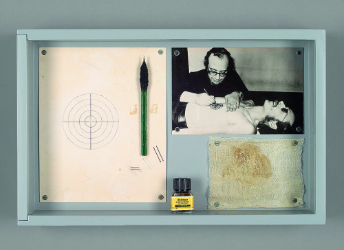 
     <i>Timm Ulrichs als menschliche Zielscheibe /Timm Ulrichs as a human target</i>, 
     1971/1974<br />
     tattooing performance and documentation material, wooden case, acrylglass window, paper, ink, tattooing needle, black white photography, muslin bandage, 
      25.5 x 40 x 7 cm<br />
     